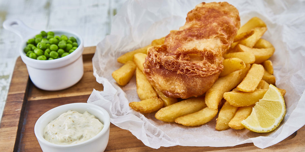 Gluten Free Beer Battered Fish served on top of chips, with a lemon wedge, peas and tartare sauce on baking parchment